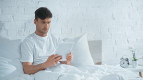 man in white t-shirt lying in bed, using digital tablet and smiling in morning