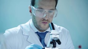 handsome scientist in protective glasses making analysis with microscope and writing down results in laboratory
