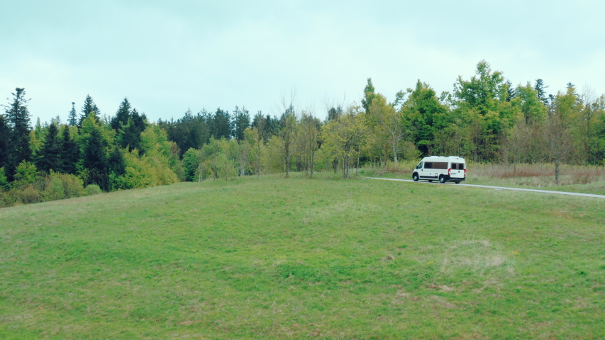 Aerial view of a white camper van driving on a small road with a view of a forest.