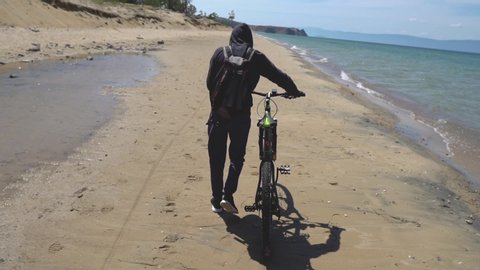 Man tourist traveler in a hoodie and backpack pushing the bike goes on a beautiful sandy beach alone. View from the back