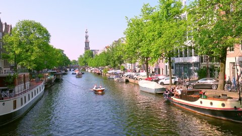 AMSTERDAM, NETHERLANDS - MAY 20, 2018: Small boats going along amazing canal, green trees at the sides. Beautiful european capital. Ancient building on a background. Sunny spring day, clear blue sky