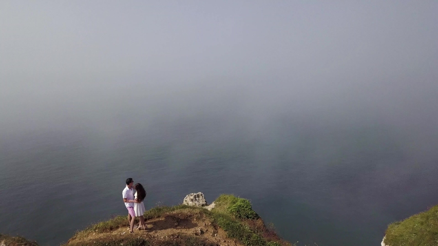 Flying over the cliff with loving couple to the blue cloudy sky above water. Action. Aerial view of seascape with the horizon hidden behind the clouds.