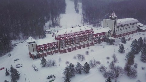 Aerial view of beautiful building among trees in mountains covered by snow. Clip. Beautiful hotel at ski resort near forested hills, sport and recreation concept.