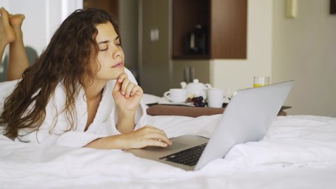 Woman have breakfast in modern hotel apartment. Window light portrait young girl in bathrobe lying on bed using laptop for work. Out of focus tray with meal, juice, plate, teapot on background