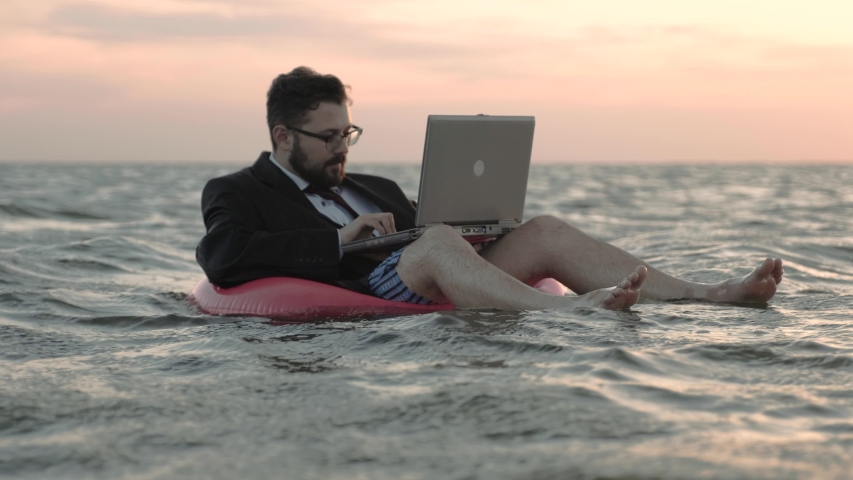 Businessman enthusiastically working behind a laptop swaying on the waves of a reservoir and sitting on a pink inflatable circle. Royalty-Free Stock Footage #1032562409