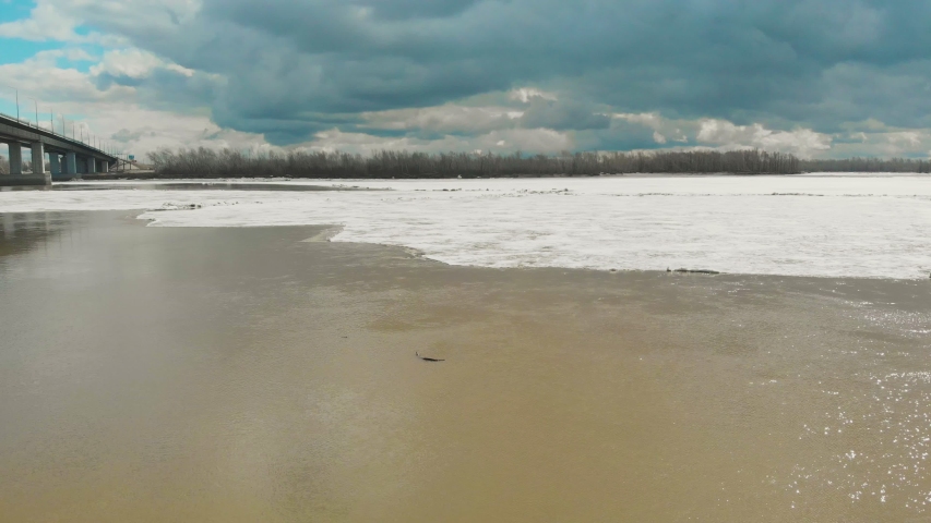 landscape of broad partly melted from ice running river with concrete bridge against bare forest and blue sky