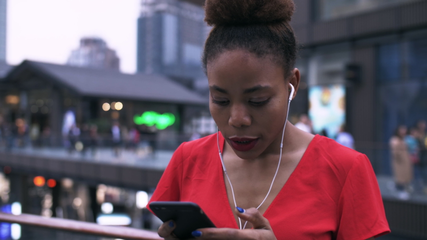Close up of One young African woman looking at mobile phone in hand and listening to music use earphone in red dress at summer urban city street of Chengdu China | Shutterstock HD Video #1032577871