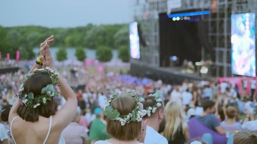 Women are watching concert at open air music festival, back view, stage lights and dancing fan crowd at background
