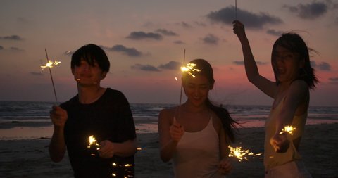 Group of Friends having fun during night party at the seaside with bengal sparkler lights in their hands. Young teenagers partying on the beach with fireworks. Slow motion shot.
