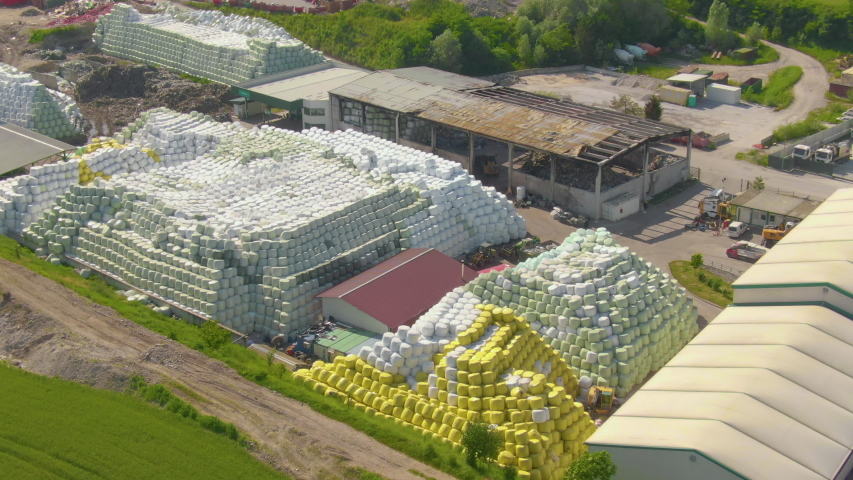 AERIAL: Flying around a large farming compound full of large heaps of hay bales wrapped in yellow and white plastic. Drone perspective of a massive farm