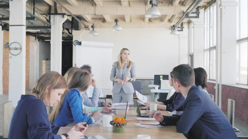 The team leader conducts a presentation near a large table and a flipchart and describes business processes. Employees sit at a table. Office life in the background. Coworking | Shutterstock HD Video #1032605489
