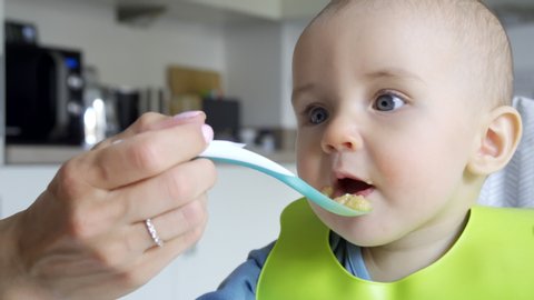 Smiling Baby Boy At Home In High Chair Being Fed Solid Food By Mother With Spoon  