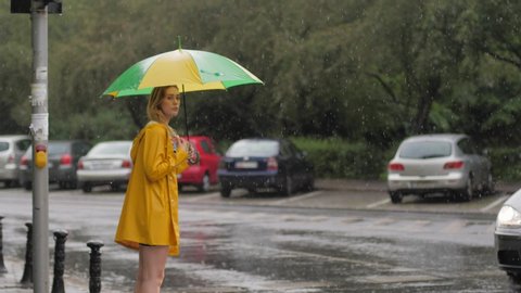 Rainy grey day lovely young woman in yellow coat hitch-hiking under shower rain with umbrella walks near traffic road in a city
