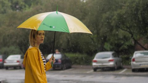 Rainy grey day lovely young woman in yellow coat hitch-hiking under shower rain with umbrella walks near traffic road in a city