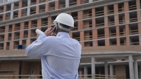 Builder in blue shirt talks on telephone on project site. Worker in white helmet talks on smartphone at construction site 4 K
