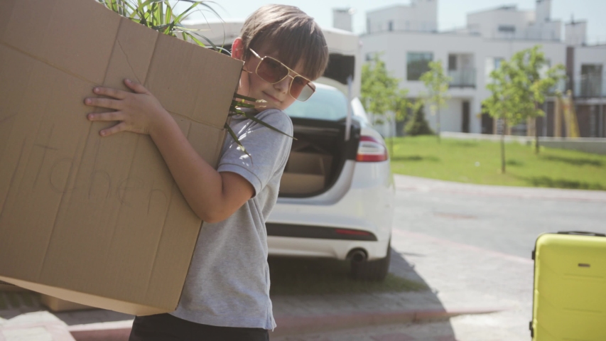 Excited young modern family moving to a new apartment in a beautiful neighbourhood. Portrait of a cute strong boy carrying big boxes to the house on sunny day.