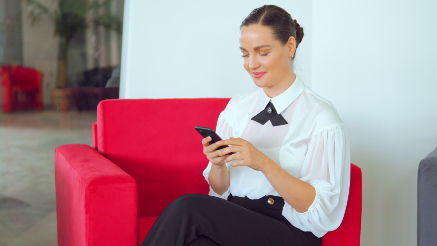 Bautiful female in white blouse is chilling in red sofa while surfing in smartphone. She seems to be happy and cheerful