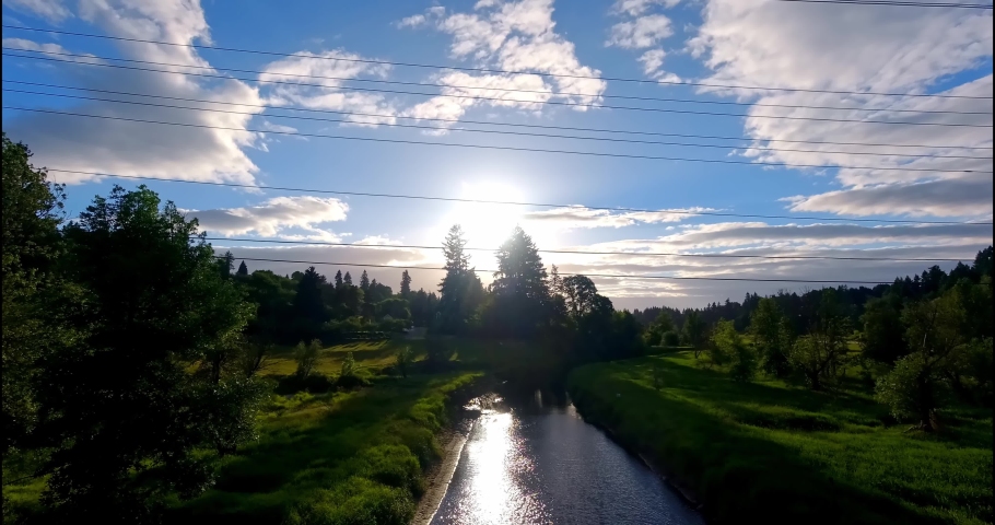 Blue sky with passing clouds reflect on a fast moving river below, surrounded by lush greenery and several evergreen trees. The sun peeks through the clouds before dusk.