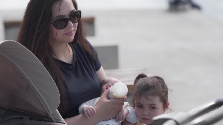 Young beautiful mother is feeding her little baby with a bottle of a milk. Her older daughter is playing near her.