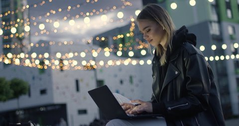 Young girl in wireless headphones uses a laptop sitting outside in a modern residential area, evening in the city, blurry lights on background, 4k slow motion.