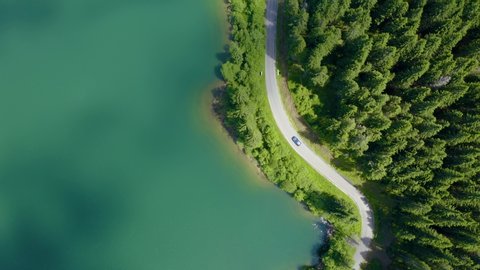 Aerial view of car driving through the forest and the lake on the side. Beautiful mountain road. Driving on the mountain road. Road trip. Mountain forest lake landscape Car driving through pine forest