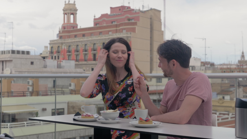 A couple is sitting in an outside cafe on the top of the building in Valencia. There are some delicious looking desserts in front of them. The guy is treating his girlfriend with an ice cream