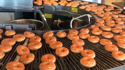 Freshly baked with a warm sugar glaze donuts going round on a conveyor belt waiting to be boxed by the baker. Filmed in 4k at cinematic 24 fps.