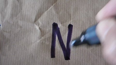 hand writes the alphabetical capital block letter N on wrapping paper using a black felt tip pen
