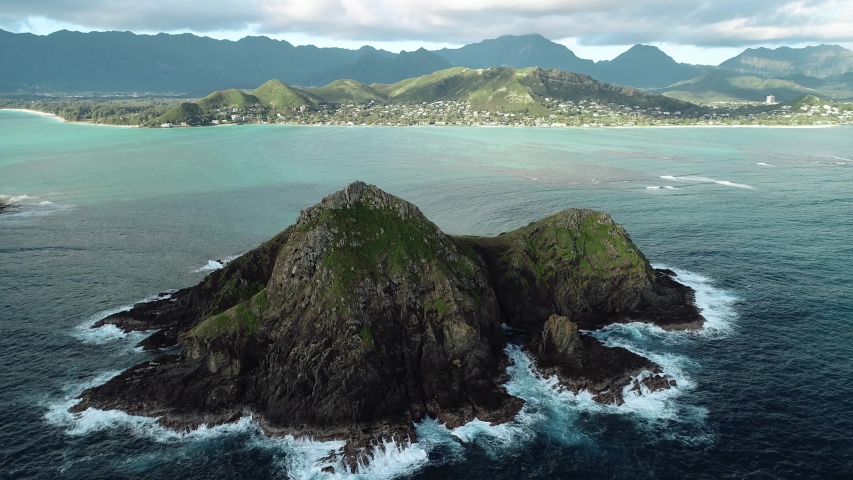 Aerial view of Lanikai beach, Honolulu, Hawaii, low angle view with static drone camera, waves breaking on the rocks of green island in the middle of the bay, color graded