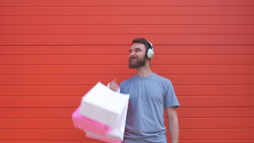 Portrait of a positive hipster beard man holding pink and white shopping bag on red background. Man listening to music in a retro headphone