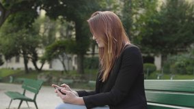 Attractive business woman using smartphone laughing Sitting on bench in park in Paris, in Horizontal Landscape Mode, watching movie, Watching Content, Videos, Red hair. Happy girl.