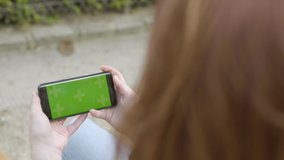 Woman using smartphone with Green Mock-up Screen Sitting on bench in park, in Horizontal Landscape Mode, watching movie, Touching touch screen. Tracking Markers. Watching Content, Videos, Red hair.