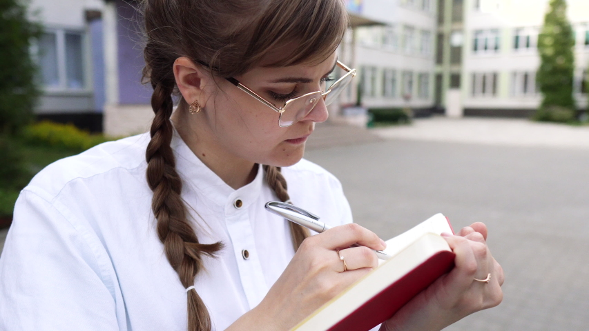 Portrait of a cute young woman in glasses and with long fair hair.?arefully writes something in a notebook.
She is a strict teacher or a smart student or a qualified doctor, schoolgirl, graduate stud