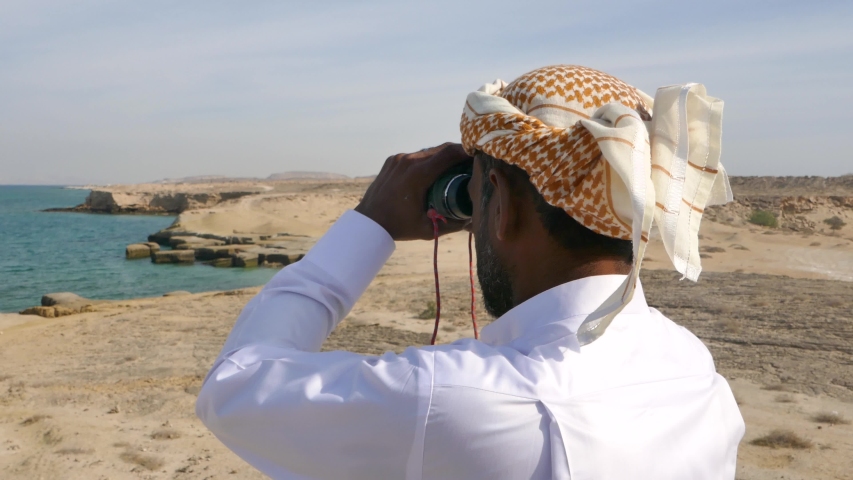 Rear back shot of an Arab man standing on a hill looking at the sea with his binoculars