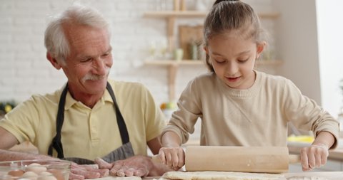 Caucasian grandpa and his granddaughter are making cookie dough. little girl playfully touches nose with her floured finger and happily laughs - family time, family bond concept 4k