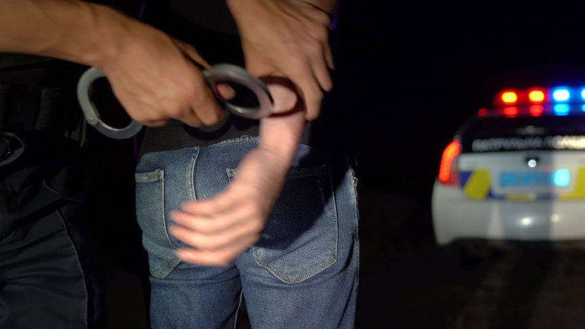 Police officer puts handcuffs on arrested man at night and leads him to a police car