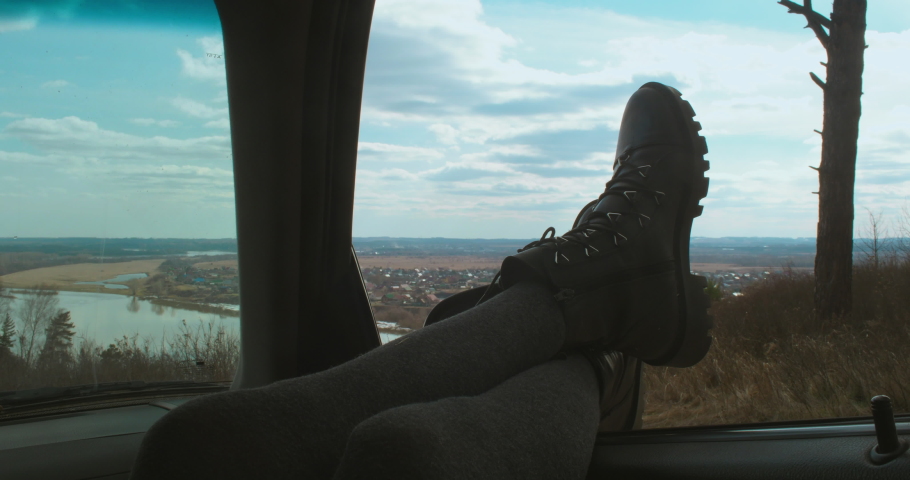 In nature, a girl in tights and black shoes, sticking her legs out of a car window, enjoys beautiful views.