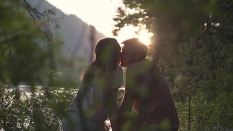 The couple in love is watching the sunset by the mountain river. A couple sits on a bench, they laugh and flirt with each other. Romantic kiss at sunset in the summer in a hike