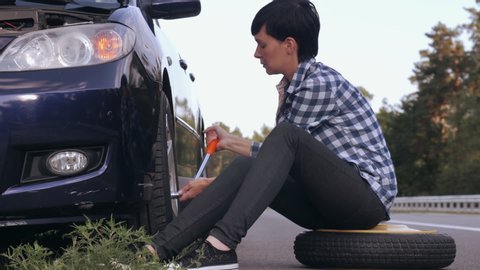 Young woman stuck on the side of the road with a flat tire. Caucasian brunette driver change a tire at the roadside.