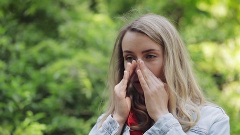 Young woman sneezes and rubs her nose standing on the park background. Allergic rhinitis person. Allergy concept.