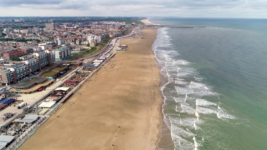 Still drone view of beach and European cityscape with blue waves crashing against yellow sand. The Hague, Netherlands