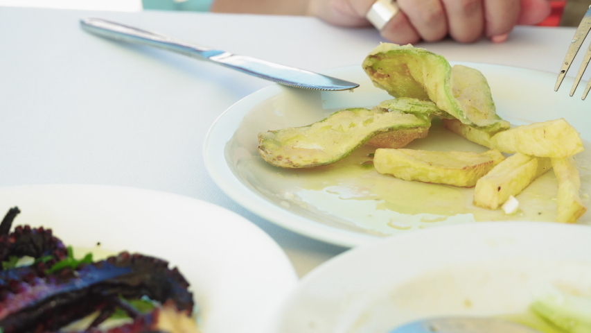 Greek tavern restaurant octopus serving closeup.
Hand with fork and knife serving crispy charred octopus tentacle on a white dish with sliced tomato, cucumber and zucchini, on a tavern table.