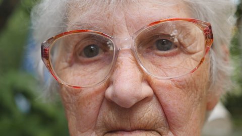 Old woman in glasses looking into camera outdoor. Portrait of sad grandmother with emotions and feelings. Granny wearing eyeglasses outside. Close up Slow motion