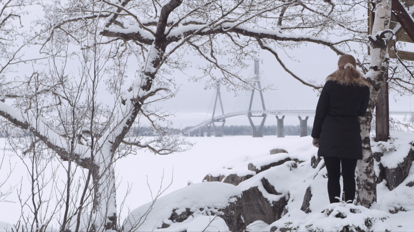 Wide Shot of a blonde woman waiting near a tree someone while looking at Replot Bridge in the distance. Vaasa (Finland) Shot on Red Monster 8K. In 4K