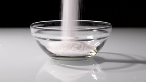 Slow-motion shot of some sugar being poured into a small glass bowl with a dark background.