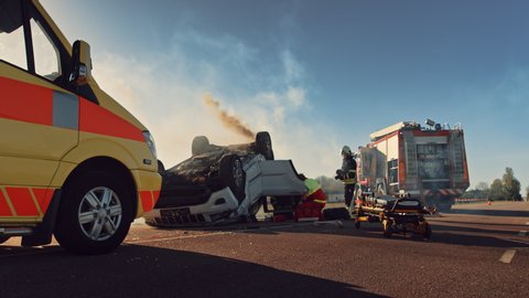 Paramedics and Firefighters Arrive On the Car Crash Traffic Accident Scene. Professionals Rescue Injured Victim Trapped in Rollover Vehicle by Extricating Them, giving First Aid and Extinguishing Fire