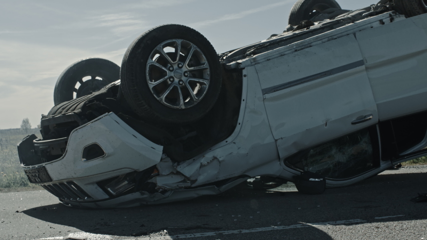 Horrific Traffic Accident Rollover Smoking and Burning Vehicle Lying on its Roof in the Middle of the Road after Collision. Daytime Crash Scene with Damaged Car. Low Angle Low Contrast Shot Royalty-Free Stock Footage #1032836240