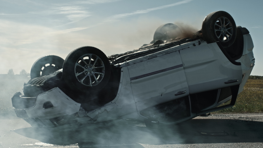 Horrific Traffic Accident Rollover Smoking and Burning Vehicle Lying on its Roof in the Middle of the Road after Collision. Daytime Crash Scene with Damaged Car. Low Angle Low Contrast Shot Royalty-Free Stock Footage #1032836243
