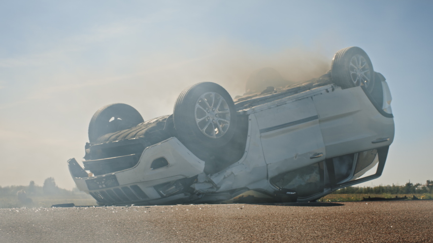 Horrific Traffic Accident Rollover Smoking and Burning Vehicle Lying on its Roof in the Middle of the Road after Collision. Daytime Crash Scene with Damaged Car. Low Angle Sunny Day Shot Royalty-Free Stock Footage #1032836252