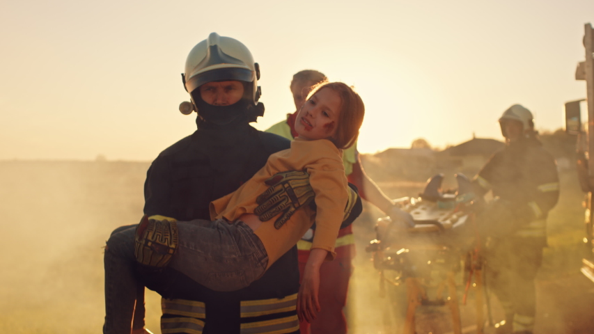 Brave Firefighter Carries Injured Young Girl to Safety. In the Background Car Crash Traffice Accident with Courageous Paramedics and Firemen Save Lifes, Fight Fire Royalty-Free Stock Footage #1032836336